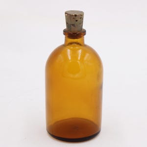 May include: Amber glass bottle with a cork stopper. The bottle has a rounded body and a narrow neck. The cork is a light brown color. The bottle is empty and sits on a white surface. A vintage apothecary bottle.