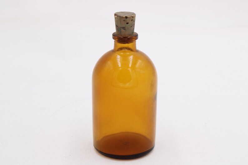 May include: Amber glass bottle with a cork stopper. The bottle has a rounded body and a narrow neck. The cork is a light brown color. The bottle is empty and sits on a white surface. A vintage apothecary bottle.