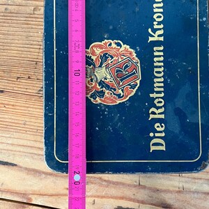 May include: A blue metal box with the text "Die Rotmann Krone" and a decorative emblem. A pink and yellow measuring tape is placed on top, showing measurements up to 30 inches.