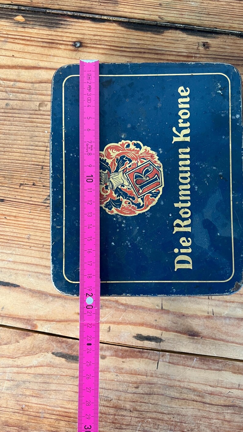 May include: A blue metal box with the text "Die Rotmann Krone" and a decorative emblem. A pink and yellow measuring tape is placed on top, showing measurements up to 30 inches.
