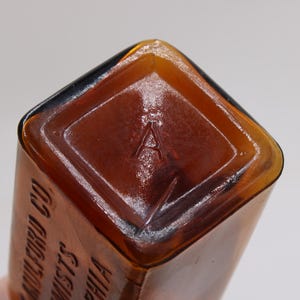 May include: Close-up of a vintage amber glass bottle. The bottom of the bottle features a diamond-shaped indentation with the letter "A" embossed in the center. The bottle's edges are rounded and the glass has a glossy finish.