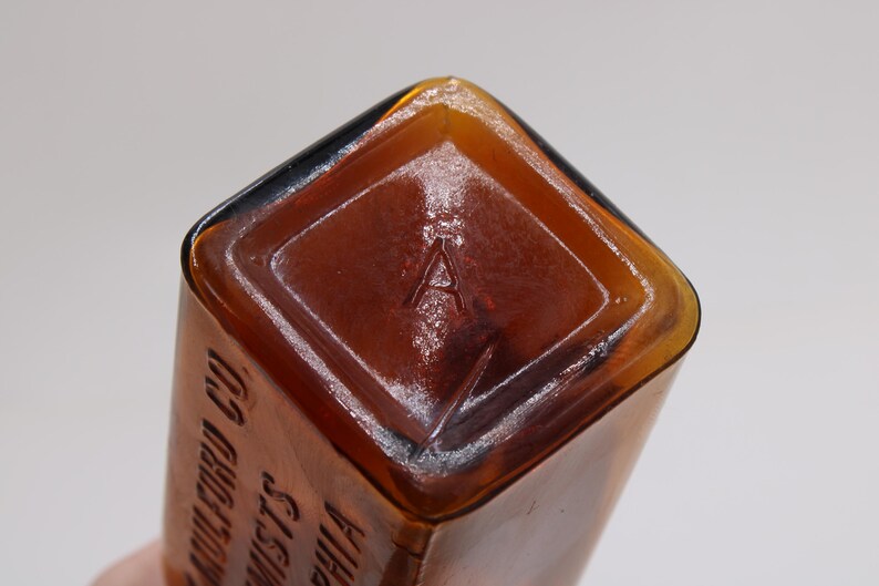 May include: Close-up of a vintage amber glass bottle. The bottom of the bottle features a diamond-shaped indentation with the letter "A" embossed in the center. The bottle's edges are rounded and the glass has a glossy finish.