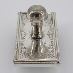 May include: A rectangular silver ink stamp with a rounded handle. The stamp's surface is decorated with floral patterns. The handle is a rounded sphere shape. The stamp is likely vintage and made of metal.