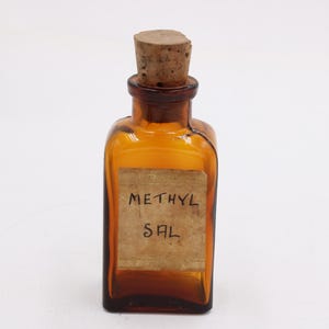 May include: An amber-colored, square glass bottle with a cork stopper. The bottle has a paper label with the handwritten text "METHYL SAL". The bottle is likely a vintage apothecary or laboratory item.