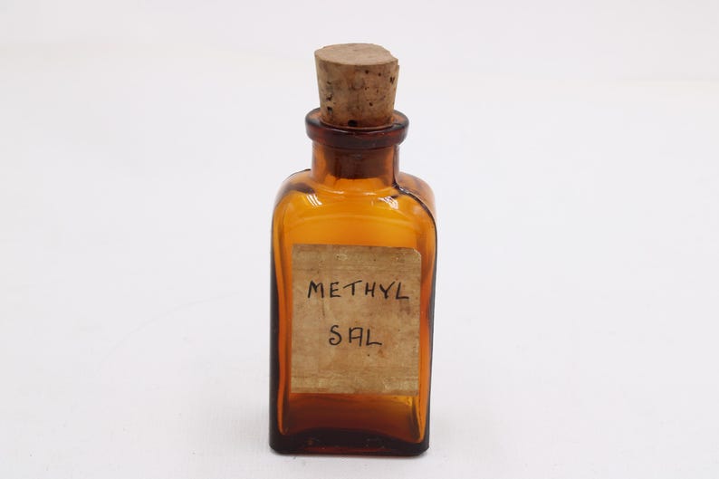 May include: An amber-colored, square glass bottle with a cork stopper. The bottle has a paper label with the handwritten text "METHYL SAL". The bottle is likely a vintage apothecary or laboratory item.