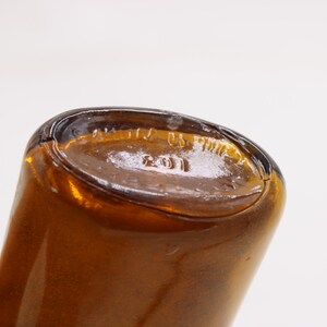 May include: Close-up of the base of a brown glass bottle. The base is oval-shaped with embossed text including the number "2011" and the words "US". The bottle has a glossy finish.
