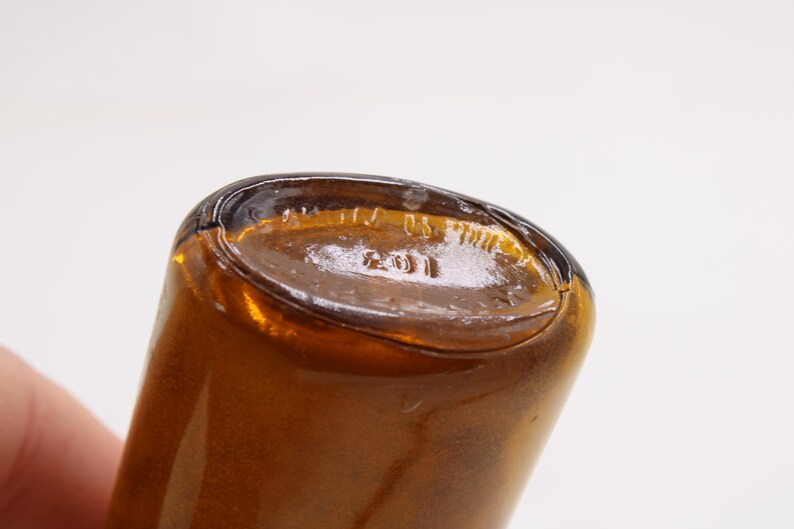 May include: Close-up of the base of a brown glass bottle. The base is oval-shaped with embossed text including the number "2011" and the words "US". The bottle has a glossy finish.