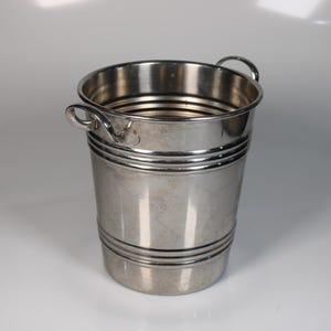 Puede incluir: Un cubo de metal plateado con dos asas pequeñas. El cubo tiene forma cilíndrica con bandas horizontales alrededor del cuerpo. El interior es visible y el cubo parece estar vacío. El fondo es una superficie blanca lisa.