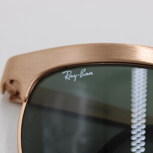 May include: Close-up of Ray-Ban sunglasses. The frame is a brushed bronze color, and the lenses are dark green. The Ray-Ban logo is visible on the lens. The sunglasses have a classic design.