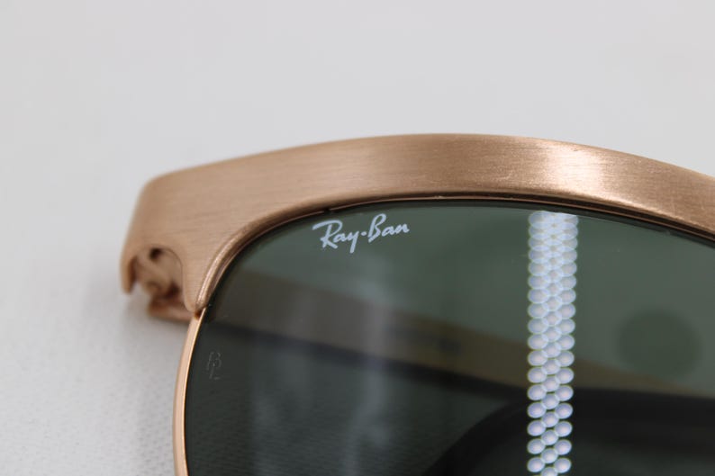 May include: Close-up of Ray-Ban sunglasses. The frame is a brushed bronze color, and the lenses are dark green. The Ray-Ban logo is visible on the lens. The sunglasses have a classic design.