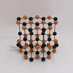 May include: A geometric model composed of black and orange spheres connected by thin rods, forming a three-dimensional cube-like structure. The spheres are evenly spaced, creating a lattice pattern. The model is likely used for educational or scientific purposes.