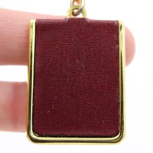 May include: A rectangular pendant with a burgundy leather-like surface, framed by a gold-colored metal border. The pendant is attached to a gold-colored chain. The background is white.