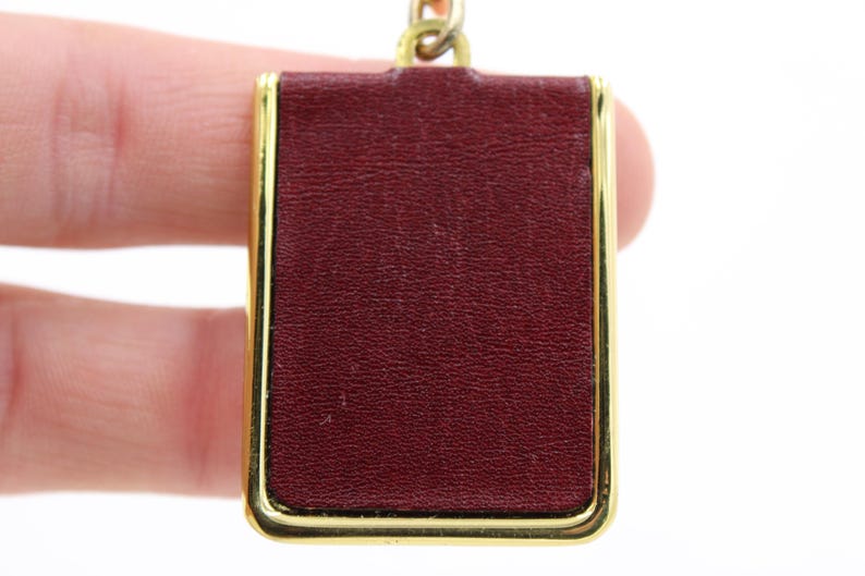 May include: A rectangular pendant with a burgundy leather-like surface, framed by a gold-colored metal border. The pendant is attached to a gold-colored chain. The background is white.