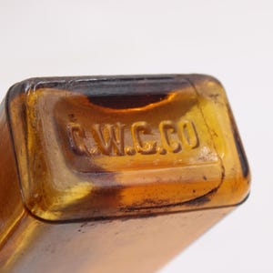 May include: Close-up of a vintage amber glass bottle with the embossed text "C.W.C.CO" on the top. The bottle has a rectangular shape and is made of a translucent, brown-tinted glass. The image highlights the details of the embossed text.