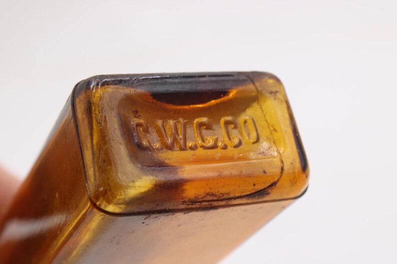 May include: Close-up of a vintage amber glass bottle with the embossed text "C.W.C.CO" on the top. The bottle has a rectangular shape and is made of a translucent, brown-tinted glass. The image highlights the details of the embossed text.