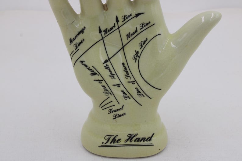May include: A pale yellow ceramic hand figurine with black lines and text indicating palmistry features. The base reads "The Hand". Lines include "Marriage Lines", "Heart Line", and "Life Line".