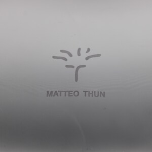 K&ouml;nnte beinhalten: Nahaufnahme einer grauen Oberfl&auml;che mit dem Namen "MATTEO THUN" in Grau gedruckt. Oberhalb des Namens befindet sich ein stilisiertes Baumdesign. Das Bild ist einfach und minimalistisch.