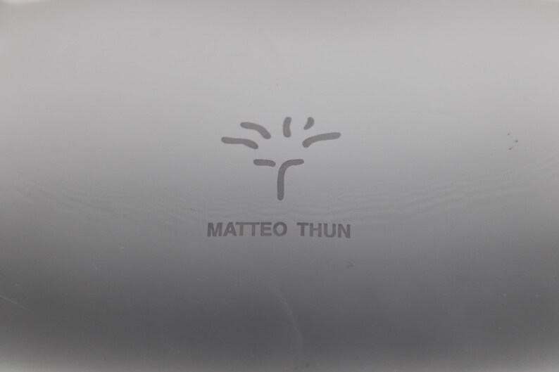 K&ouml;nnte beinhalten: Nahaufnahme einer grauen Oberfl&auml;che mit dem Namen "MATTEO THUN" in Grau gedruckt. Oberhalb des Namens befindet sich ein stilisiertes Baumdesign. Das Bild ist einfach und minimalistisch.