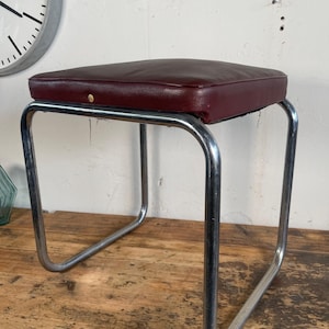 以下が含まれることがあります： バーガンディ色の革張りのシートとクロムメッキのスチールフレームを備えたヴィンテージスツール。シートは正方形でパッド入り、フレームは湾曲したチューブデザインです。ミッドセンチュリーモダン家具のクラシックな作品です。