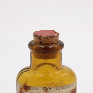 May include: An antique amber glass bottle with a cork stopper. The bottle has a label with faded text and a measurement scale. The cork is sealed with red wax. The bottle is likely from a pharmacy or laboratory.