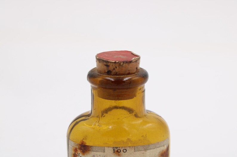 May include: An antique amber glass bottle with a cork stopper. The bottle has a label with faded text and a measurement scale. The cork is sealed with red wax. The bottle is likely from a pharmacy or laboratory.