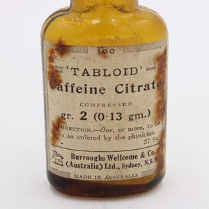May include: An old amber glass bottle with a label stating "TABLOID Caffeine Citrate." The label details the dosage, "gr. 2 (0.13 gm.)", and the manufacturer, Burroughs Wellcome & Co., Made in Australia. The bottle is a vintage pharmaceutical item.