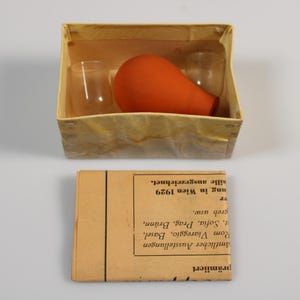 Puede incluir: Un antiguo botiquín médico con una caja que contiene dos pequeños viales de vidrio y una pera de goma naranja. Debajo del kit hay una pequeña caja rectangular con texto.