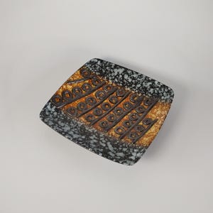 May include: A square ceramic dish with a speckled black and white border and a textured, orange and brown center. The center features a repeating pattern of brown circles and lines. The dish is likely for decorative or serving purposes.