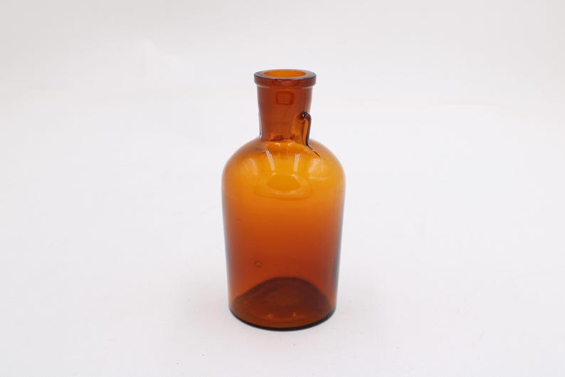 May include: Amber-colored glass bottle with a rounded body and a narrow neck. The bottle has a small handle on the side and a slightly flared lip. The glass has a smooth surface and a warm, inviting tone. The bottle is empty and stands on a white surface.