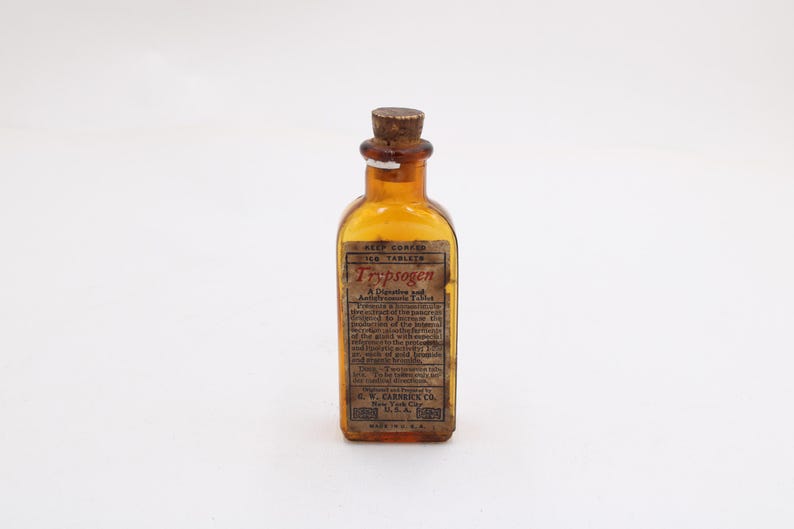 May include: An amber glass bottle with a cork stopper. The bottle features a rectangular label with the word "Trypsogen" in red. The label includes text describing the product and the manufacturer, G.W. Carnrick Co., New York, U.S.A. This is likely an antique bottle.