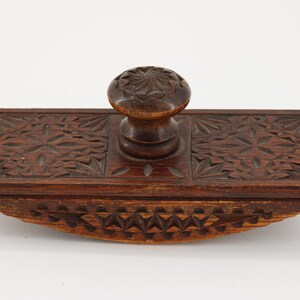 May include: A vintage wooden ink blotter with intricate carved floral and geometric patterns. The blotter features a rectangular base with a curved bottom and a central knob for handling. The wood has a rich, dark brown color.