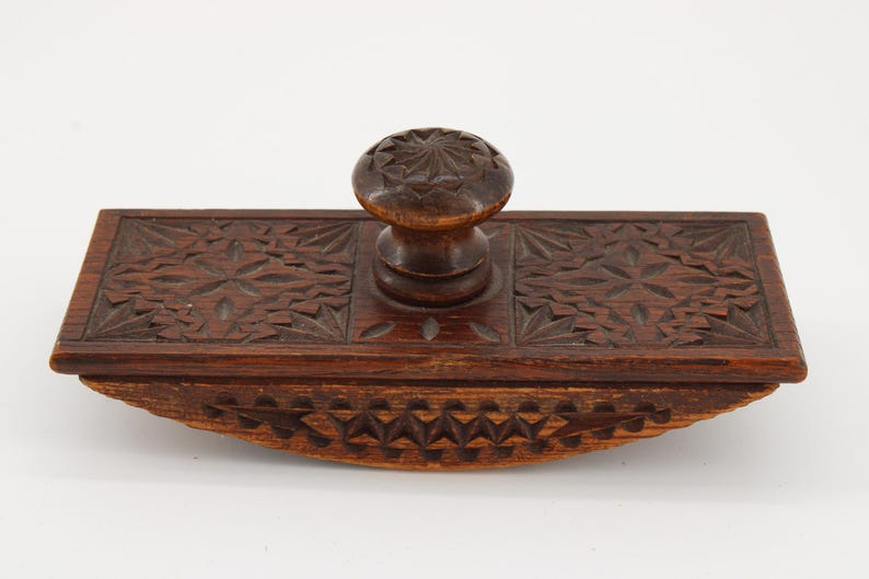 May include: A vintage wooden ink blotter with intricate carved floral and geometric patterns. The blotter features a rectangular base with a curved bottom and a central knob for handling. The wood has a rich, dark brown color.