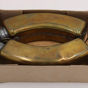 May include: Two antique brass flasks with screw-on caps, nestled in a cardboard box. The flasks have a curved, elongated shape and a tarnished, aged appearance. The box is plain, with visible wear.