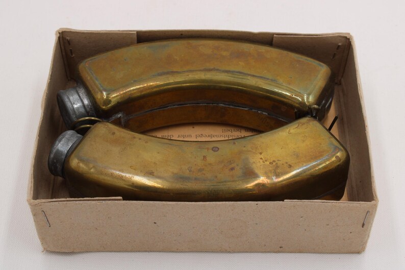May include: Two antique brass flasks with screw-on caps, nestled in a cardboard box. The flasks have a curved, elongated shape and a tarnished, aged appearance. The box is plain, with visible wear.