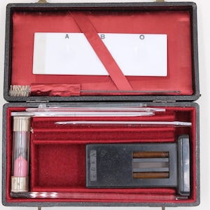 May include: Open black case with red velvet lining, containing medical instruments. Includes a white slide labeled A, B, and O, glass tubes, a brush, and a timer with pink sand. A vintage medical kit.