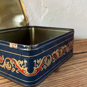 May include: A vintage rectangular tin box with a navy blue exterior. The box features a decorative gold and red ornate design around the edges. The lid is open, revealing a gold interior.