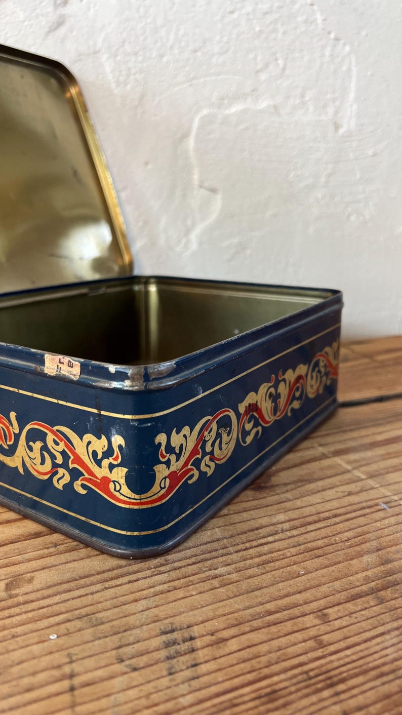 May include: A vintage rectangular tin box with a navy blue exterior. The box features a decorative gold and red ornate design around the edges. The lid is open, revealing a gold interior.