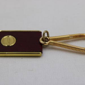 May include: A rectangular keychain with a burgundy leather surface and a gold-colored border. A gold emblem is centered on the leather. The keychain has a gold-colored, coiled wire loop for attaching keys.