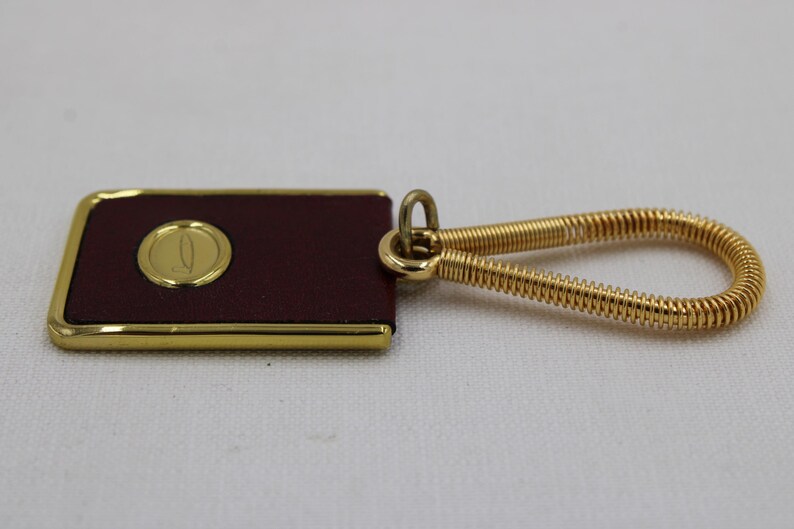 May include: A rectangular keychain with a burgundy leather surface and a gold-colored border. A gold emblem is centered on the leather. The keychain has a gold-colored, coiled wire loop for attaching keys.