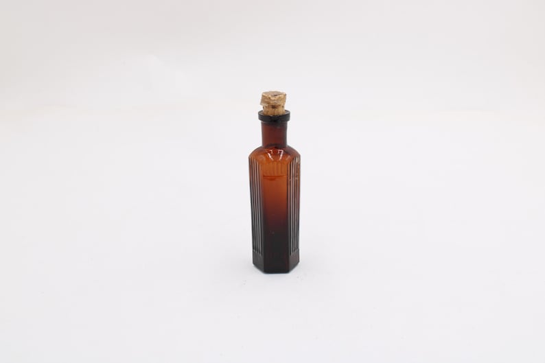 Puede incluir: Una peque&ntilde;a botella de vidrio marr&oacute;n antiguo con tap&oacute;n de corcho. La botella tiene forma rectangular con estr&iacute;as verticales y un color &aacute;mbar oscuro. El corcho es marr&oacute;n claro y texturizado.