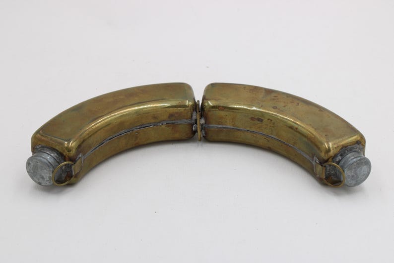 May include: Two-part brass flask with a curved, banana-like shape. Each half has a screw-on cap made of a silver-colored metal. The flask is designed to be separated into two pieces. The brass has a slightly aged appearance.