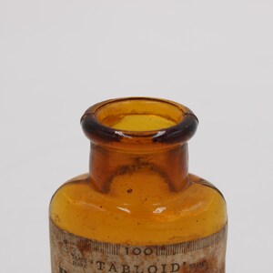 Puede incluir: Primer plano de una botella de vidrio &aacute;mbar vintage con un borde marr&oacute;n oscuro. La botella tiene forma cil&iacute;ndrica y una etiqueta con la palabra "TABLOID" visible. La botella es probablemente de principios del siglo XX.