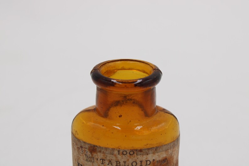 Puede incluir: Primer plano de una botella de vidrio &aacute;mbar vintage con un borde marr&oacute;n oscuro. La botella tiene forma cil&iacute;ndrica y una etiqueta con la palabra "TABLOID" visible. La botella es probablemente de principios del siglo XX.