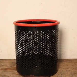 Puede incluir: Papelera de metal negro con borde rojo. La papelera tiene un diseño perforado.