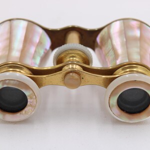 May include: Antique opera glasses with a gold-tone metal frame and iridescent mother-of-pearl shell casing. The lenses are black, and the focus wheel is white. A vintage accessory.