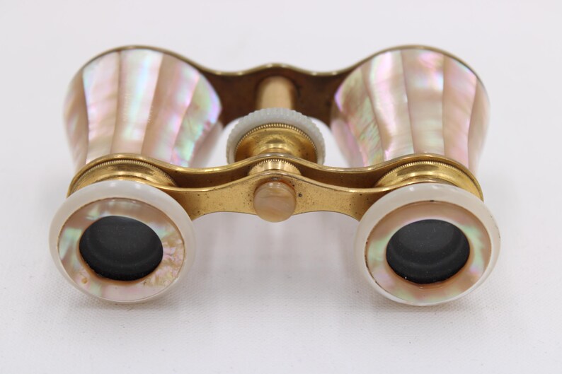 May include: Antique opera glasses with a gold-tone metal frame and iridescent mother-of-pearl shell casing. The lenses are black, and the focus wheel is white. A vintage accessory.