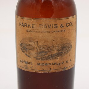 Puede incluir: Una botella de vidrio marr&oacute;n vintage con una etiqueta de papel. La etiqueta presenta el texto "PARKE, DAVIS & CO. MANUFACTURING CHEMISTS" y una ilustraci&oacute;n de un edificio. La botella est&aacute; etiquetada como "DETROIT, MICHIGAN, U.S.A."