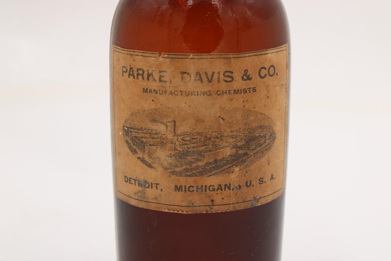 Puede incluir: Una botella de vidrio marr&oacute;n vintage con una etiqueta de papel. La etiqueta presenta el texto "PARKE, DAVIS & CO. MANUFACTURING CHEMISTS" y una ilustraci&oacute;n de un edificio. La botella est&aacute; etiquetada como "DETROIT, MICHIGAN, U.S.A."