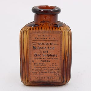 K&ouml;nnte beinhalten: Eine antike bernsteinfarbene Glasflasche mit einem Etikett mit der Aufschrift "SOLOID BRAND Boric Acid and Zinc Sulphate". Die Flasche hat ein geripptes Design und einen dunkelbraunen Deckel. Das Etikett enth&auml;lt auch das Wort "POISON".