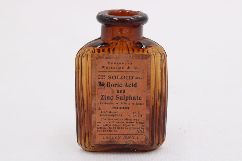 K&ouml;nnte beinhalten: Eine antike bernsteinfarbene Glasflasche mit einem Etikett mit der Aufschrift "SOLOID BRAND Boric Acid and Zinc Sulphate". Die Flasche hat ein geripptes Design und einen dunkelbraunen Deckel. Das Etikett enth&auml;lt auch das Wort "POISON".